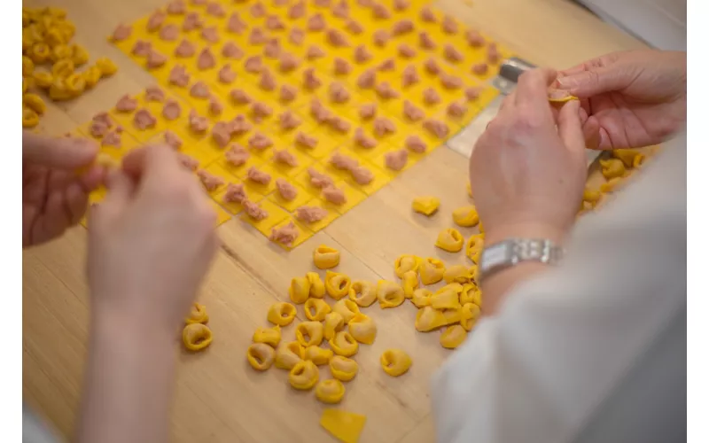 Primo piano delle mani di due persone che preparano dei tortellini, sullo sfondo il tavolo con file di tortellini da chiudere e alcuni tortellini già pronti