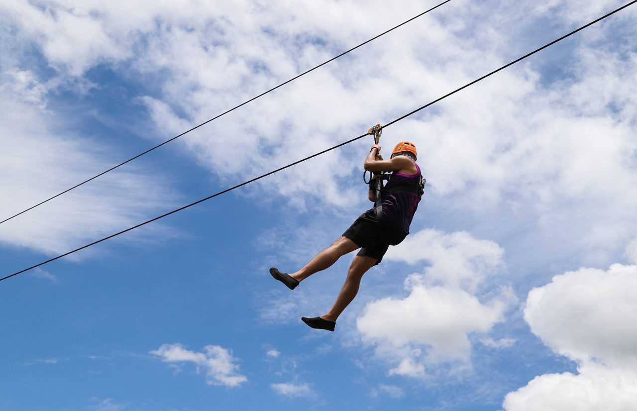 Closeup of a man gliding on extreme trolley zipline in adventure park with cloudy blue sky background in sunny day.
