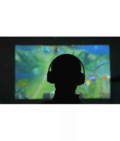 Silhouette di spalle di un uomo con delle cuffie che gioca con un videogame davanti a un grande schermo in una stanza scura