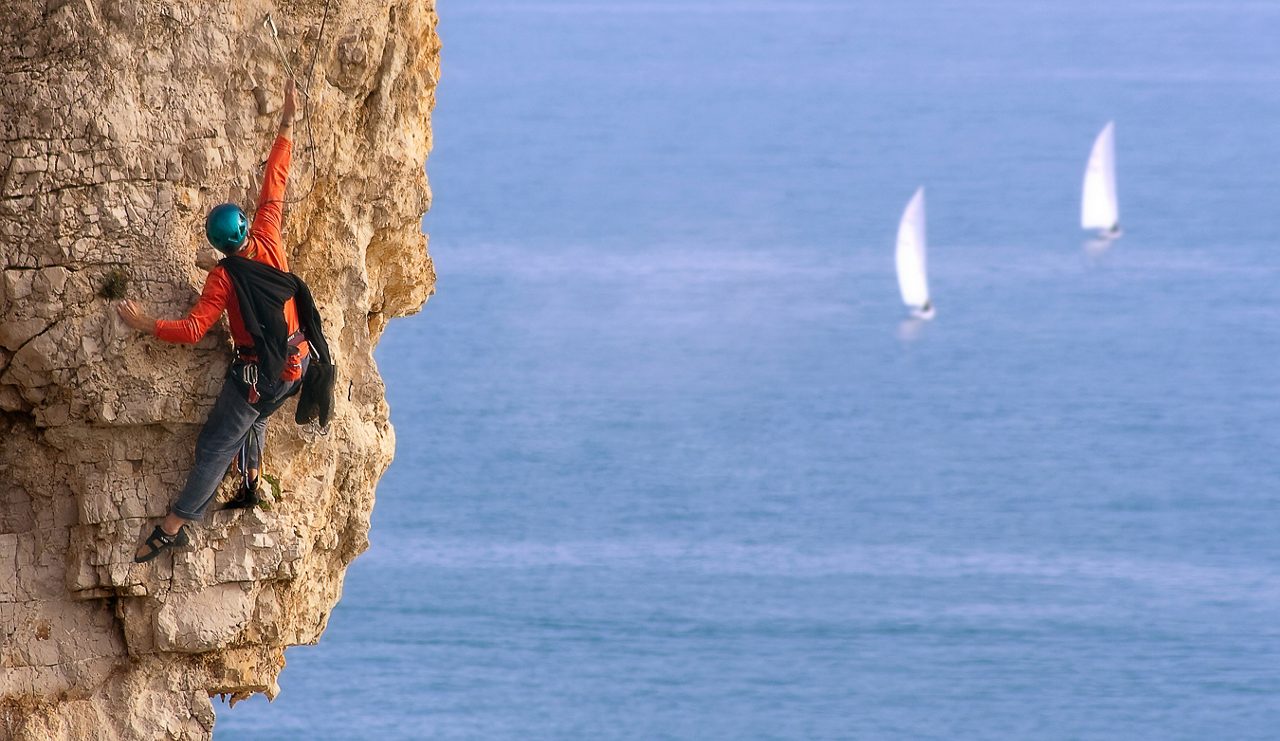 A climber on a rocky wall with two boat with sails racing in the sea below