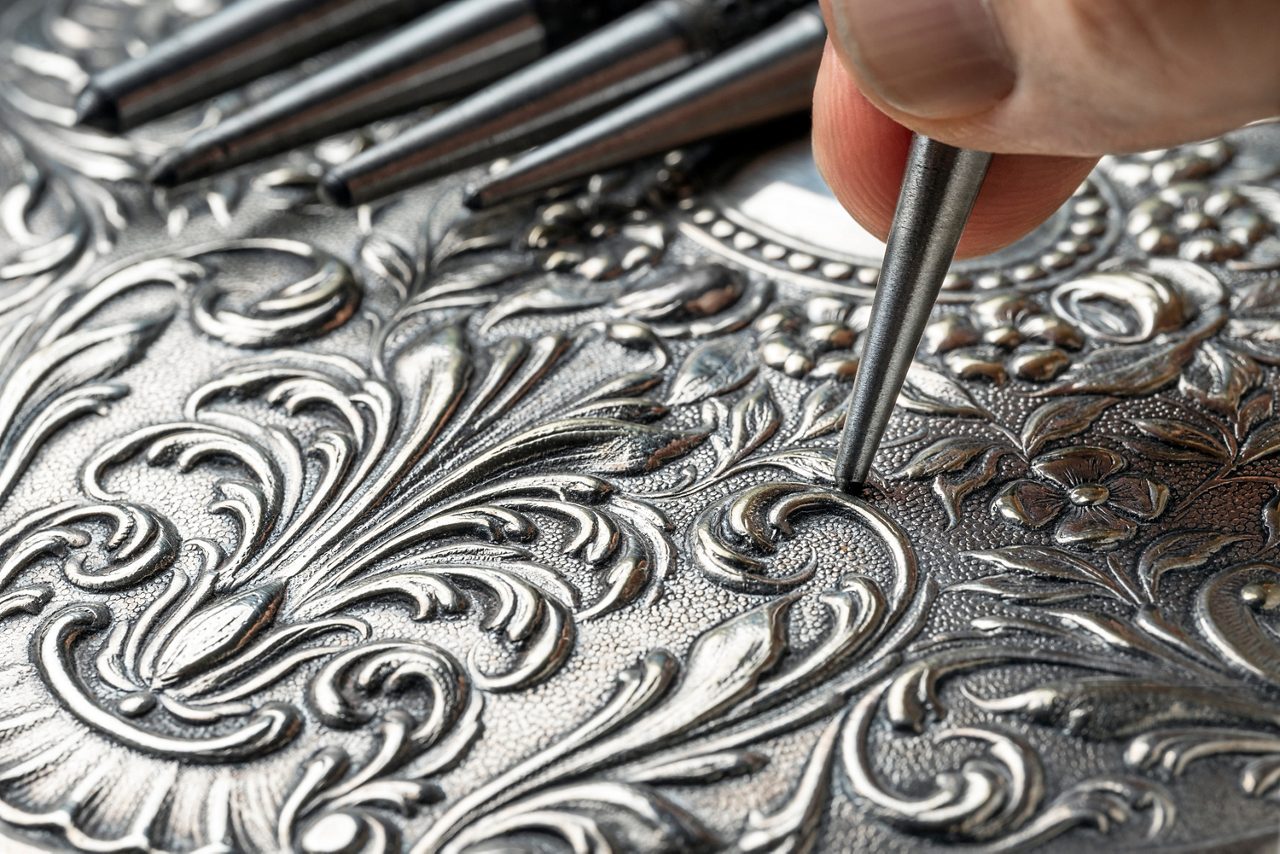 CLOSEUP OF CRAFTSMAN'S HAND EMBOSSING METAL WITH PUNCH IN THE WORKSHOP. GOLDSMITH, SILVERSMITH, JEWELLERY AND HANDICRAFTS CONCEPT.
